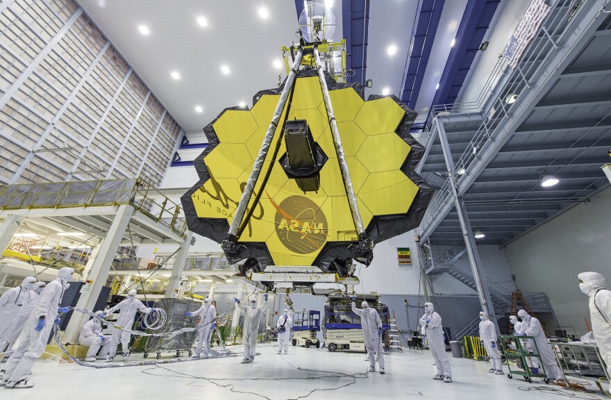 The mirror of the James Webb telescope in a large warehouse room is a series of yellow hexagons that form a circular shape. People in full white protective suits work on the floor below it.