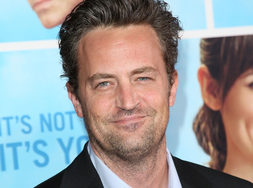 A man with slicked back brown hair, blue eyes and stubble on his face is smiling at the camera.
