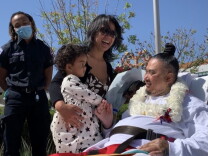 An Asian man in a stretcher smiles as a two-year-old girl is lifted up by her mother to greet him.