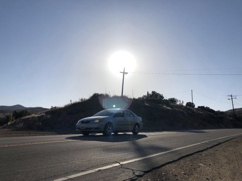 A utility pole and cross arm holding electrical lines is backlit by the sun as a car drives in the foreground