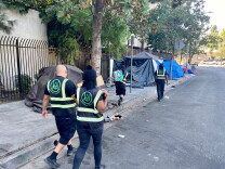 Four outreach workers from Urban Alchemy are walking down the street wearing all black and vests that identify them as part of Urban Alchemy, the nonprofit hired by Mitch O'Farrell's district to assist LAHSA with outreach. They are handing out food and water to unhoused people. 