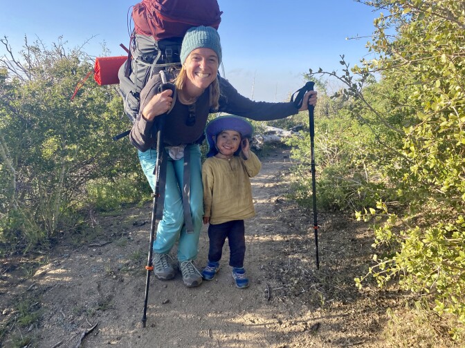 A woman, smiling and holding hiking poles, half squats next to a small boy in the middle of a trail.