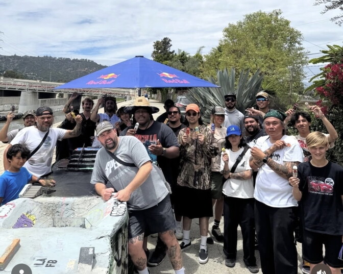 A group picture at the LA River Fingerboard Skatepark. About 20 people pose and hold up their fingerboards right next to the skatepark. A blue umbrella with a "Red Bull" logo covers some of them. A bridge is seen in the background on the left side. Shrubbery is seen on the right side.