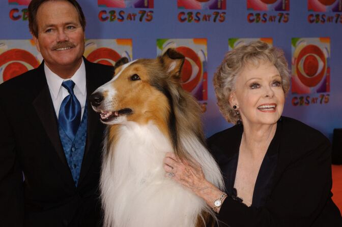 An older woman holds a collie dog next to a man with a mustache.