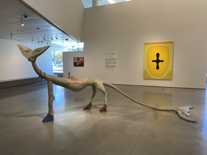 A sculpture of a mythical animal with three legs and a dolphin or whale tale stands in the middle of a gallery. A bright yellow painting is on the back wall. 