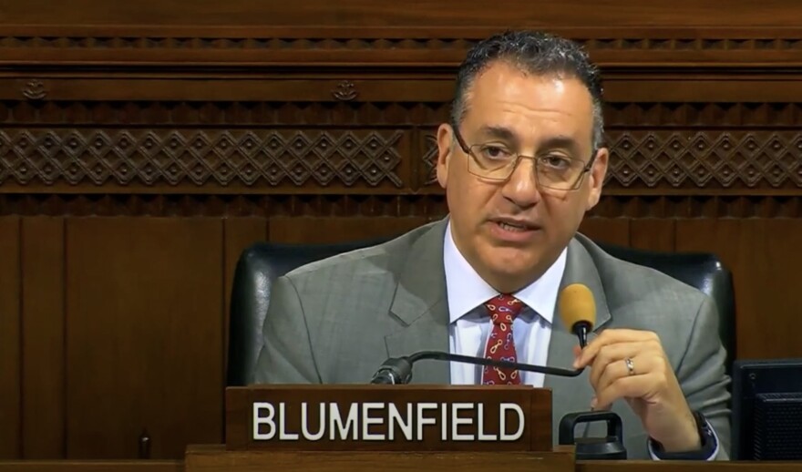 A man in a suit speaks into a microphone with the words "BLUMENFIELD" written on a name placard in front of him.