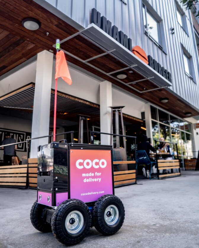 a black, box-shaped robot with four wheels and a pink and purple sign on the side that reads, "coco, made for delivery," sits outside a restaurant.