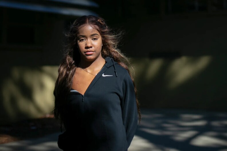 A Black female college student poses for a photo for CalMatters. She is wearing a black zip-up nike hoodie and has long brown hair. She is standing outside with a blurred background.