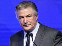 A close up shot of a clean cut, male presenting person in a dark suit speaking into a microphone in front of a blue background
