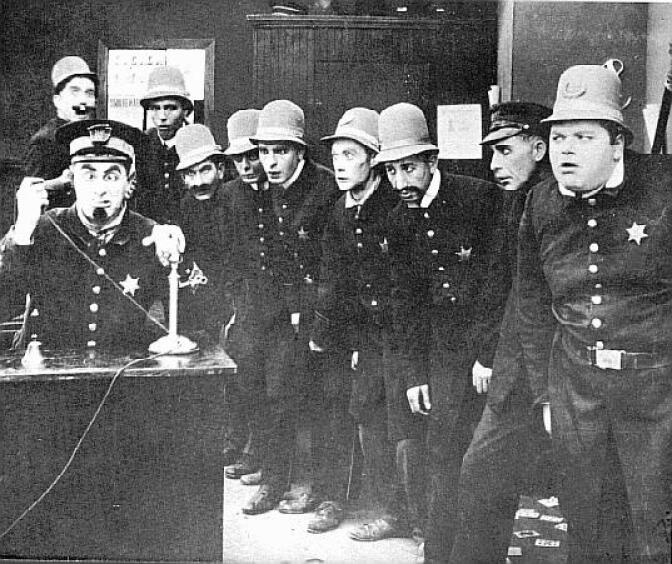 Scene from the Keytstone Cops film "Clutches of the Gang" directed by George Nichols and Mack Sennett. The desk officer using the telephone is Ford Sterling. The policeman directly behind Sterling (in extreme background, left) is Edgar Kennedy. The hefty policeman at extreme right is Roscoe "Fatty" Arbuckle. The young constable with bulging eyes, fourth from right, is Arbuckle's nephew Al St. John. The casting of the Keystone police force changed from one film to the next; many of the individual members were per-diem actors who remain unidentifiable.
