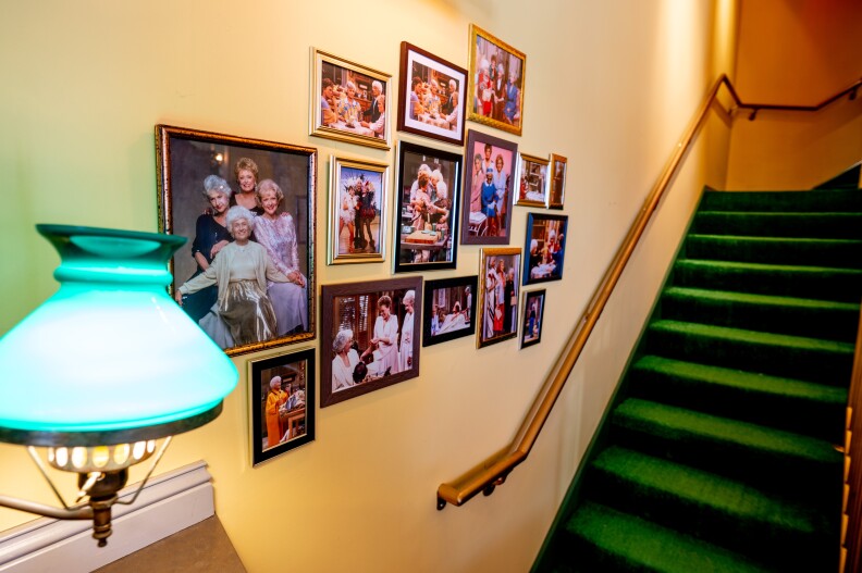 Framed photos of the Golden Girls hang on the wall alongside a green staircase and a lamp with a green shade. Pictured are Dorothy, Blanche, Rose, and Sophia, both in a posed photo and in shots from a variety of episodes of the series.