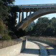 A concrete arched bridge goes over a channelized stream. Lush trees are on either side. 