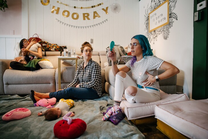 A woman with curled turquoise hair holds up a round blue plastic device in her right hand and holds her left hand to her breast. Another woman with a black and white checked shirt sits to her right and another sits on a couch with an infant in her arms.