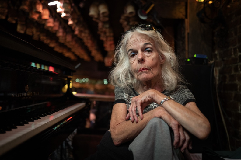 An older white woman with light skin sits next to the keys of a piano, looking at the camera.