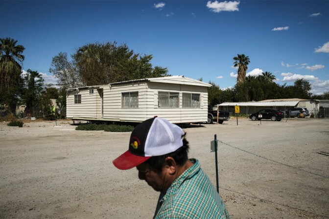 A white mobile home is shown in the middle of a dirt and gravel lot. A man with medium brown skin is shown blurred in the foreground. His face is turned away from the camera. 