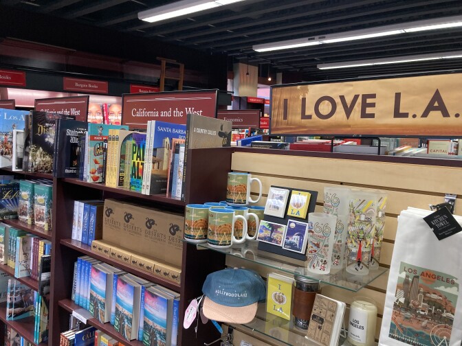 A bookstore shelf with mugs, a hat, glasses, magnets, tea towels and books. There are three rectangular signs along the top of the shelf. Two read "California and the West" and one reads "I LOVE L.A."