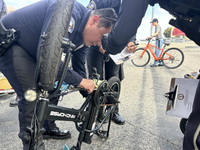 Police officers examine a folded electric bike's gears in a parking lot as another writes on a clipboard.