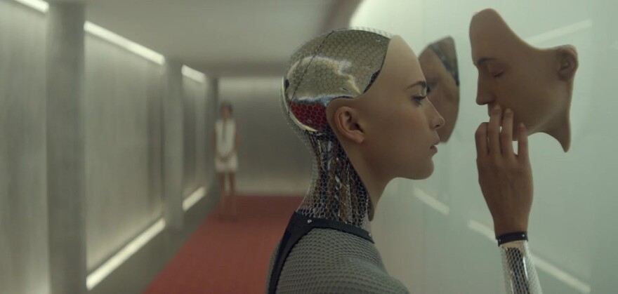 In a scene from the 2015 film "Ex Machina," a female robot reaches with her right hand to touch a robotic face hanging from a wall.