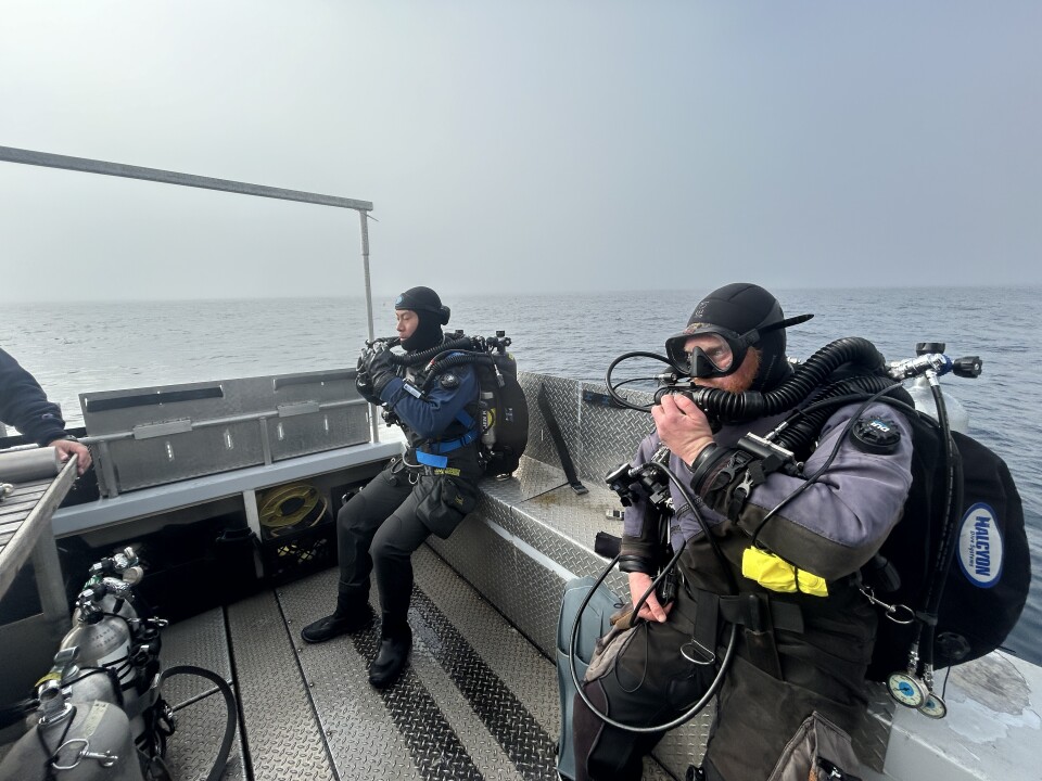 Two men sit on a deck of a boat wearing scuba diving gear. It's foggy and calm on the water. 