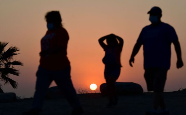Three people are silhouetted against a sunset. They all wear masks, the red sun behind them, walking down a dimly lit dusty trail with some plants visible behind them. They appear to be an adult female, a younger female, and an adult male.