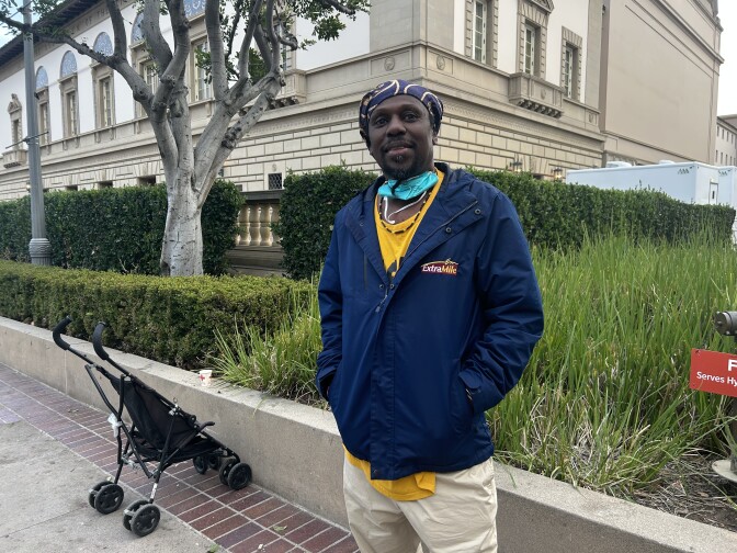 A dark-skinned man in a jacket and head covering stand outside in a courtyard.