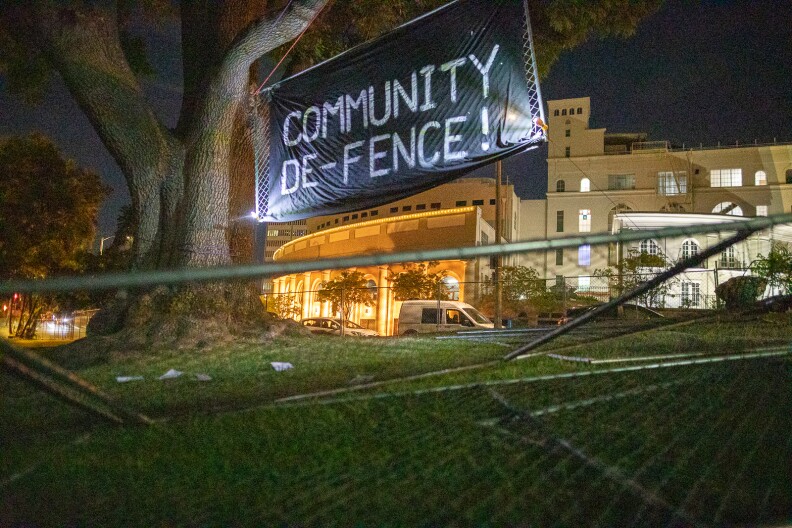 A black tarp placed over some metal fencing, hung from a tree, reads "COMMUNITY DE-FENCE!" Toppled fencing is seen in the foreground, buildings lit up in the background.