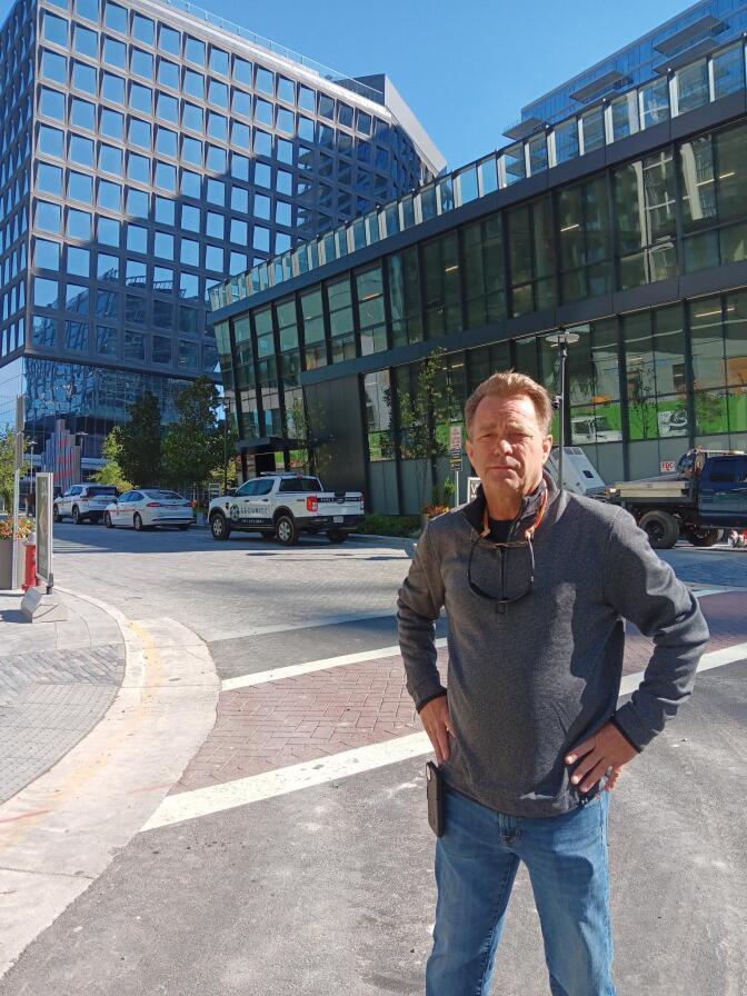 A man with light skin tone, wearing a gray quarter zip sweater and light denim pants, stands with his hands on a his waist on a street corner across from a couple buildings with large glass windows.
