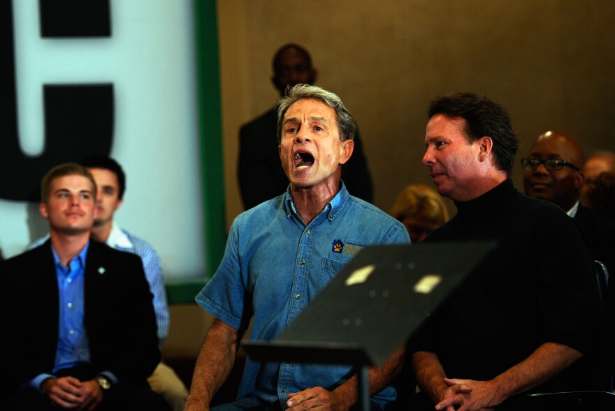 A man with short gray hair and wearing a blue denim shirt has his mouth open wide as if shouting. Other people stand or sit nearby.