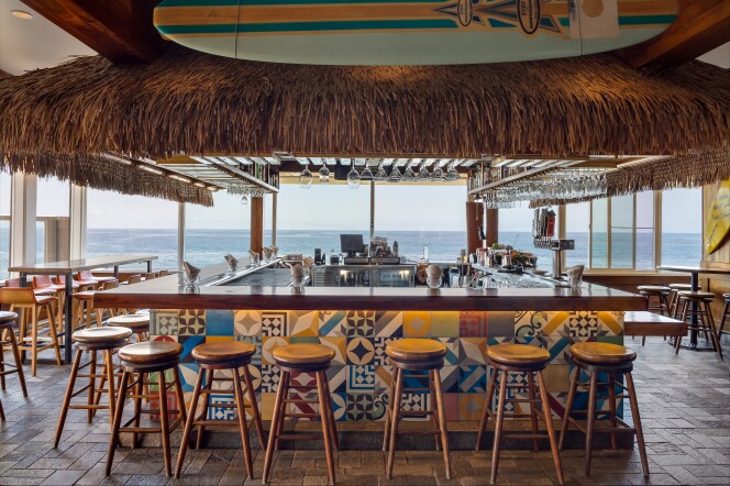 A bar overlooking the Pacific ocean: There are wooden stools lining the bar and high top tables, all positioned for a view of the waves. The decor is a little bit tiki (there's a grass fringe skirt rings the cabinetry above the bar), but also a little beachy (there's are surfboards on the wall and above the bar) and colorful tiles accent the front of the bar.