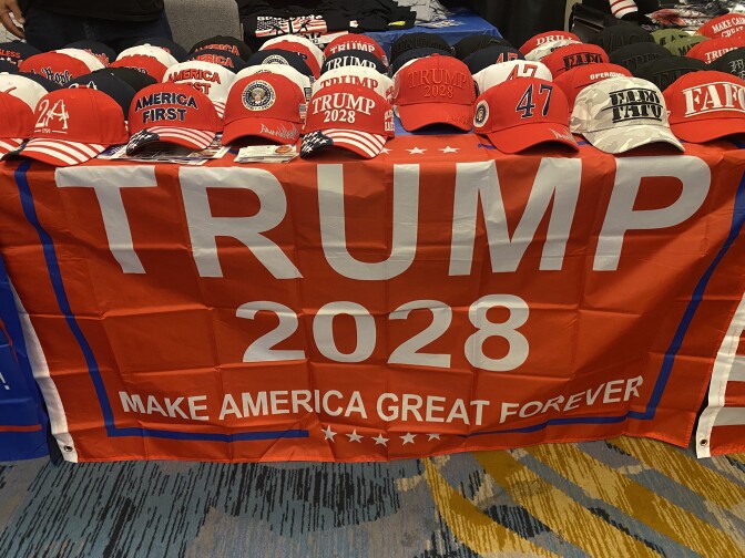 A bright red banner saying Trump 2028 hangs on a table with Trump hats for sale on top.