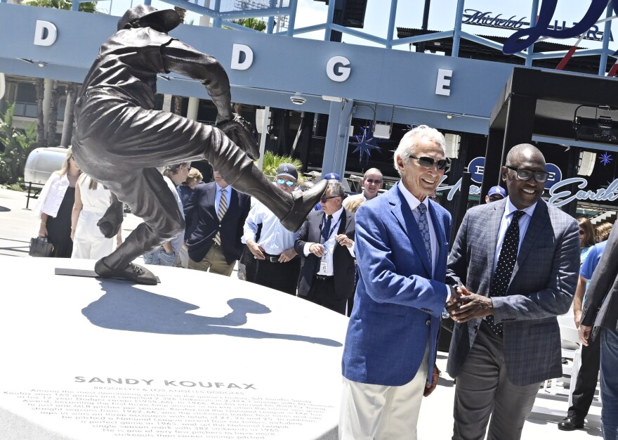 Sandy Koufax in a Dodger blue suit jacket shakes hands with a man in a dark suit and tie in front of his bronze statue.