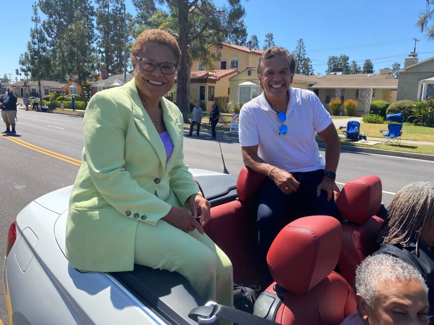 Karen Bass sits on the back of a white convertible car wearing a light green suit, a purple shirt, and glasses next to Antonio Villaraigosa who wears a white polo shirt and dark pants.