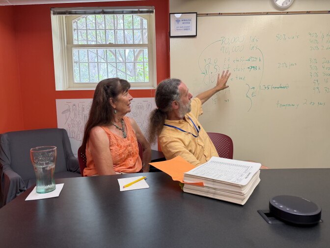 A woman in an orange sleeveless shirt next to a man in a yellow shirt. They sit at a black table top, and the man is gesturing toward writing on a white board. 