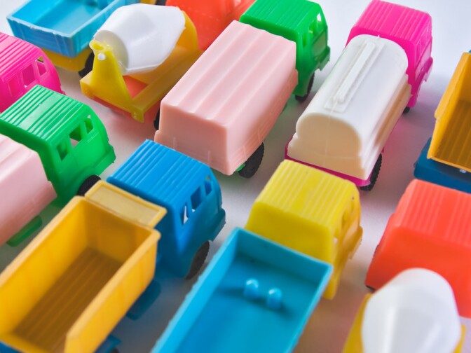 Several colorful toy trucks designed in a very simple manner, including a tank truck, a concrete transport, a refrigerator and a dump truck. Their colors are either very intense or very light. The cars are parked next to each other in two rows.
