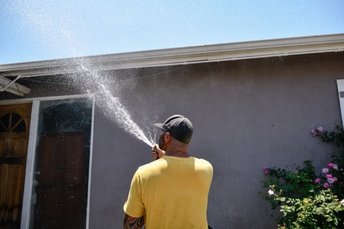 A man in a yellow T-shirt and dark gray ball cap sprays at hose toward a house.
