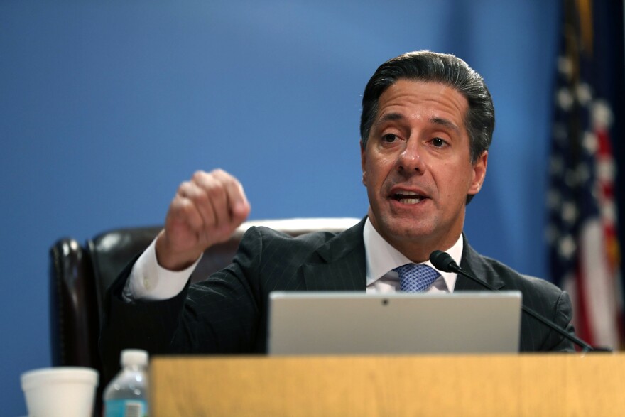 A man wearing a black suit and a tie seated at a school board dais gestures with his closed right hand while speaking into a microphone.