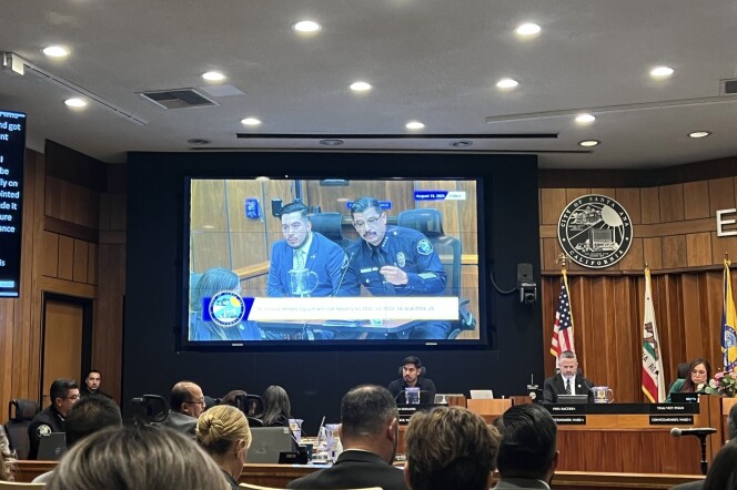 A crowded city council meeting room: Several elected officials are sitting on a dais, with flags hanging behind them. There is a screen showing two individuals speaking to the city council, one is wearing a police uniform.