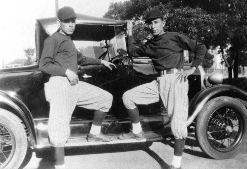 two men in baseball uniforms stand next to a vintage auto