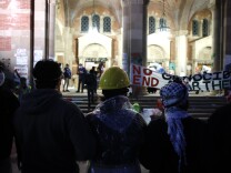 A row of people face a lighted building. A sign on a long piece of cloth reads: No Genocide