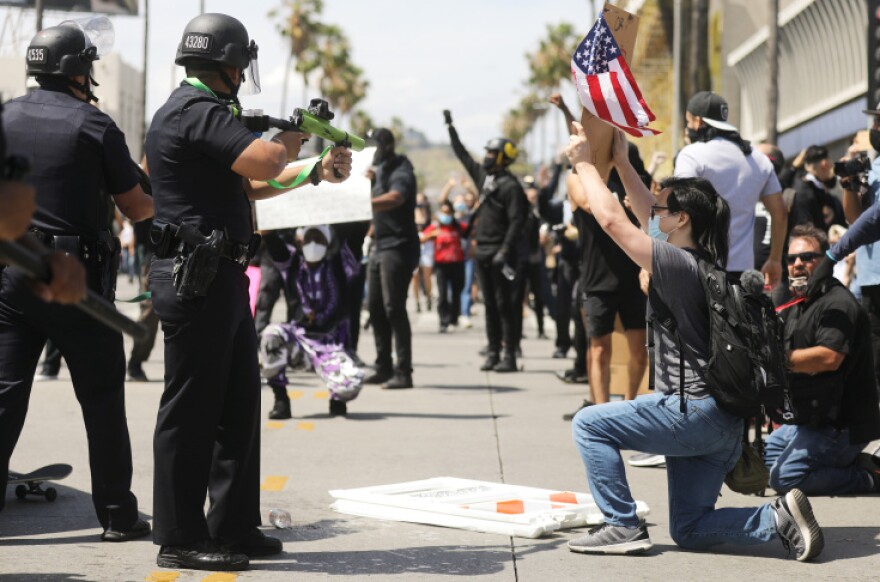 Two police officers wearing black helmets square off against protesters on a sidewalk. One holds a green and black non-lethal gun with a shoulder stock. One protester wearing a backpack has taken a knee and is holding a sign up while facing in the direction of the officers.