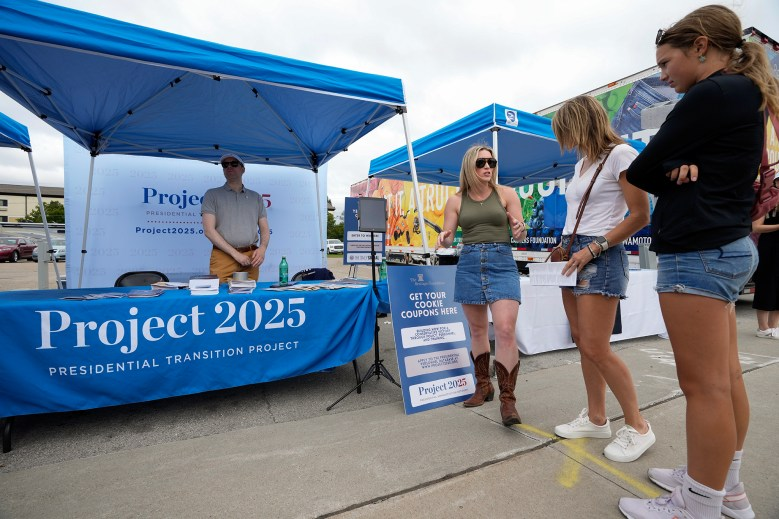 Outside there is a tent and table with a person standing behind the table's sign that reads "Project 2025"