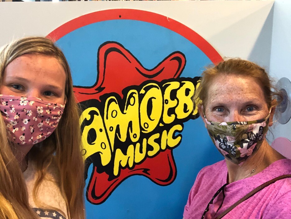 Two women, wearing masks, stand on either side of the Amoeba Music logo, which has bright yellow bubbly lettering in front of a red star shape and a blue circle.