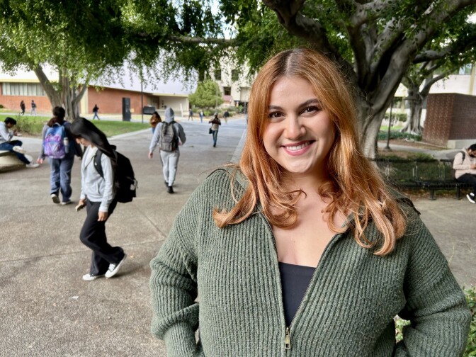 Person with shoulder length hair stands in front of a tree and smiles.