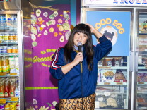 An Asian woman with dark brown hair stands in front of refrigerator doors inside a grocery store, holding a microphone, wearing a blue sports jacket. In the background are coolers filled with frozen foods, sodas, and a sign that reads El Sereno Green Grocer.