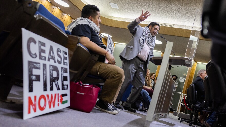 A masculine presenting person wearing a gray suit and Kaffiyeh around his neck with short dark hair and glasses speaks at a podium surrounded by people sitting in blue chairs. At the bottom left of frame is a white sign that reads "Cease Fire Now!"