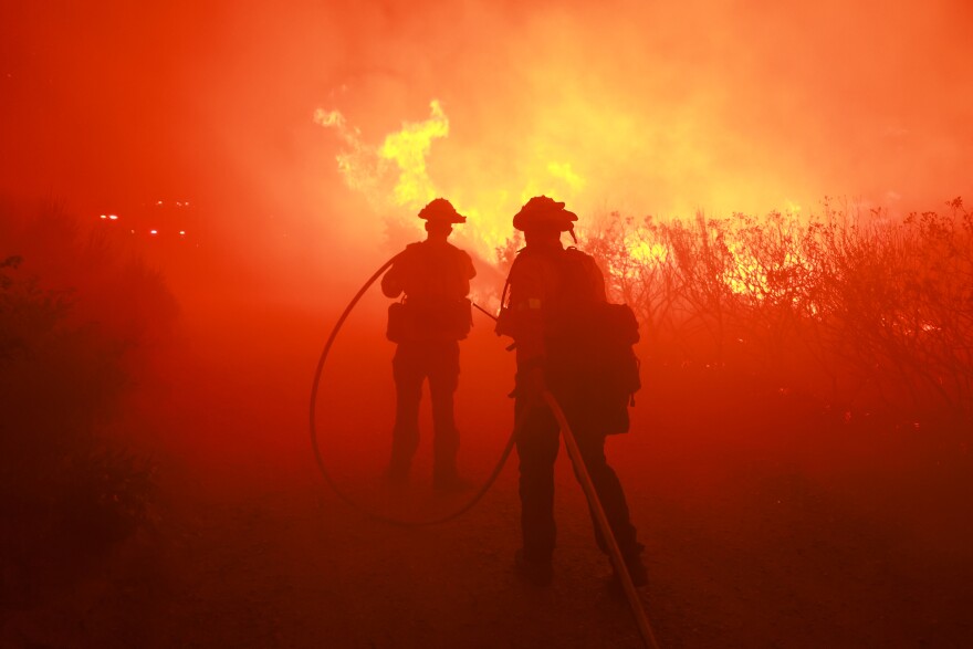 The silhouettes of firefighters in helmets and with a hose are dark agains a fire that fills the background in orange and red