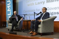 Two men sit on upholstered chairs in suits with mics in front of them. They're on a stage.