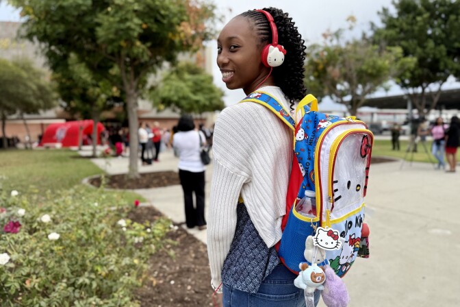 A teenage girl with dark skin tone and long curly black hair turns for a portrait outside. On her back is a blue, white, yellow and red Hello Kitty backpack. Over her ears she wears Hello Kitty headphones.