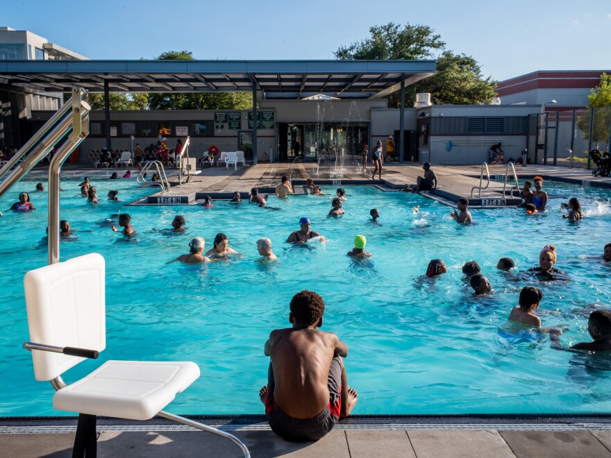 A child with brown skin tone sits at the edge of a pool with his back turned to the camera while the pool is filled with other people swimming.