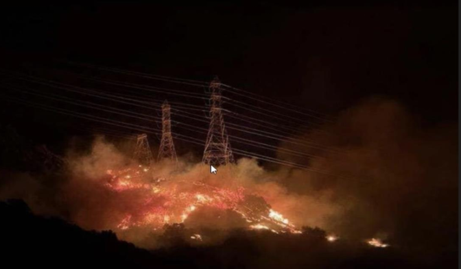 A burning hillside with powerlines within the flames is seen with smoke rising into the night sky.